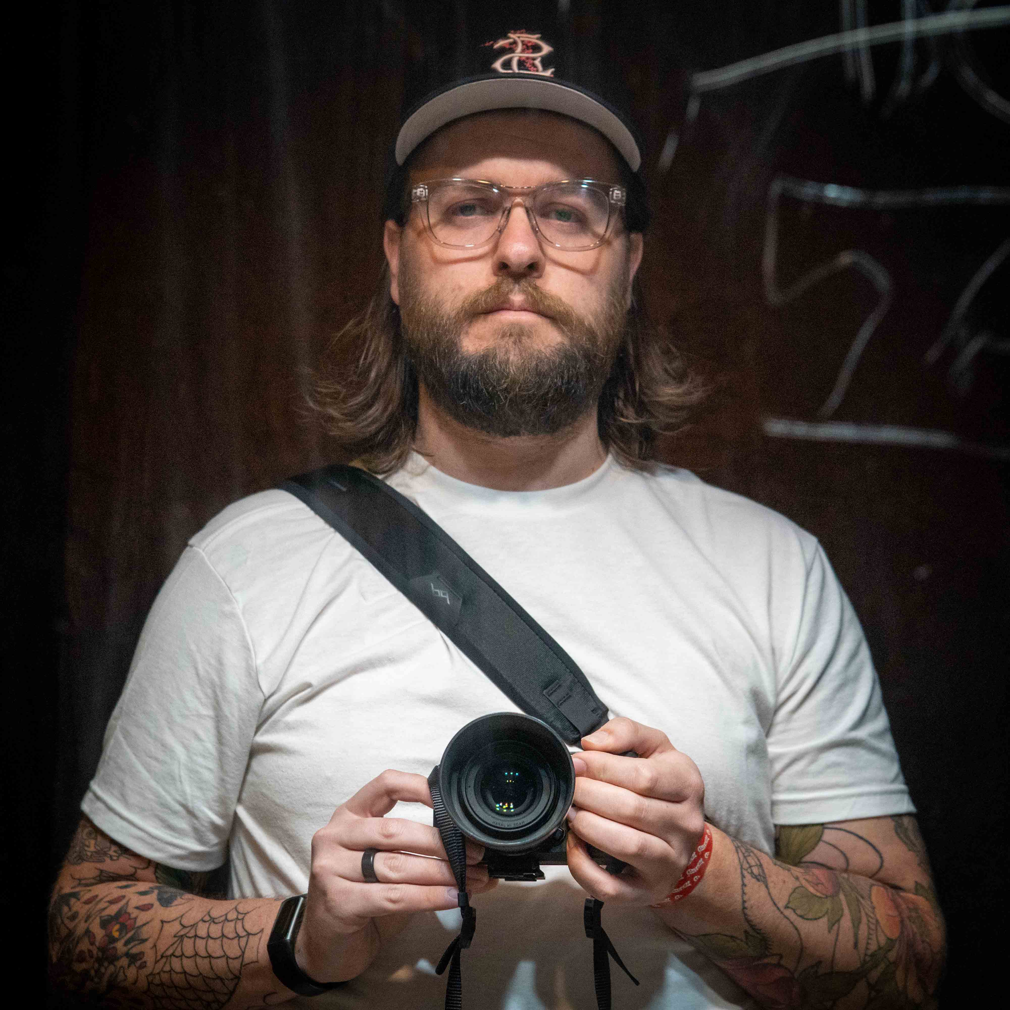 Ethan wearing clear, thick-framed glasses, a black hat, and white shirt looking into a scratched mirror while holding a camera.